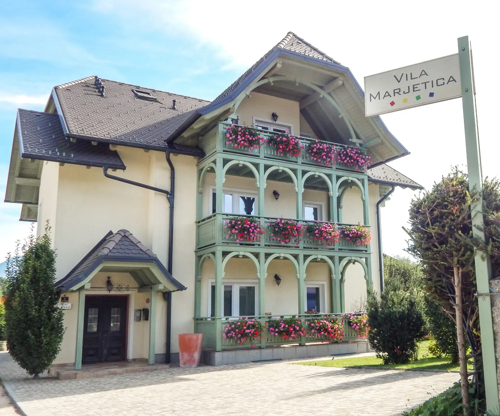 Vila Marjetica s pročelja: zeleni rezljani balkoni s pelargonijami nad vhodom